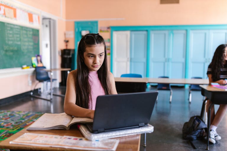A focused young girl using a laptop for schoolwork in a bright classroom environment.
