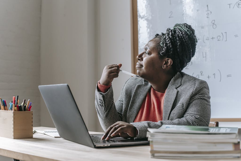 Thoughtful African American female teacher in formal outfit pondering while sitting at table and working on laptop