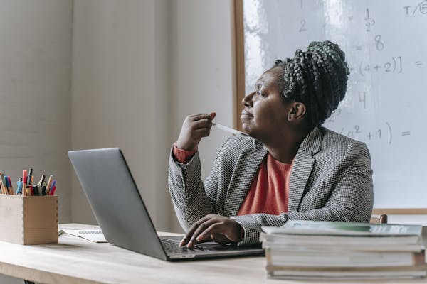 Thoughtful African American female teacher in formal outfit pondering while sitting at table and working on laptop