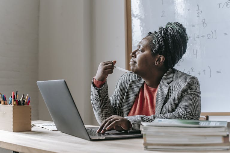 Thoughtful African American female teacher in formal outfit pondering while sitting at table and working on laptop