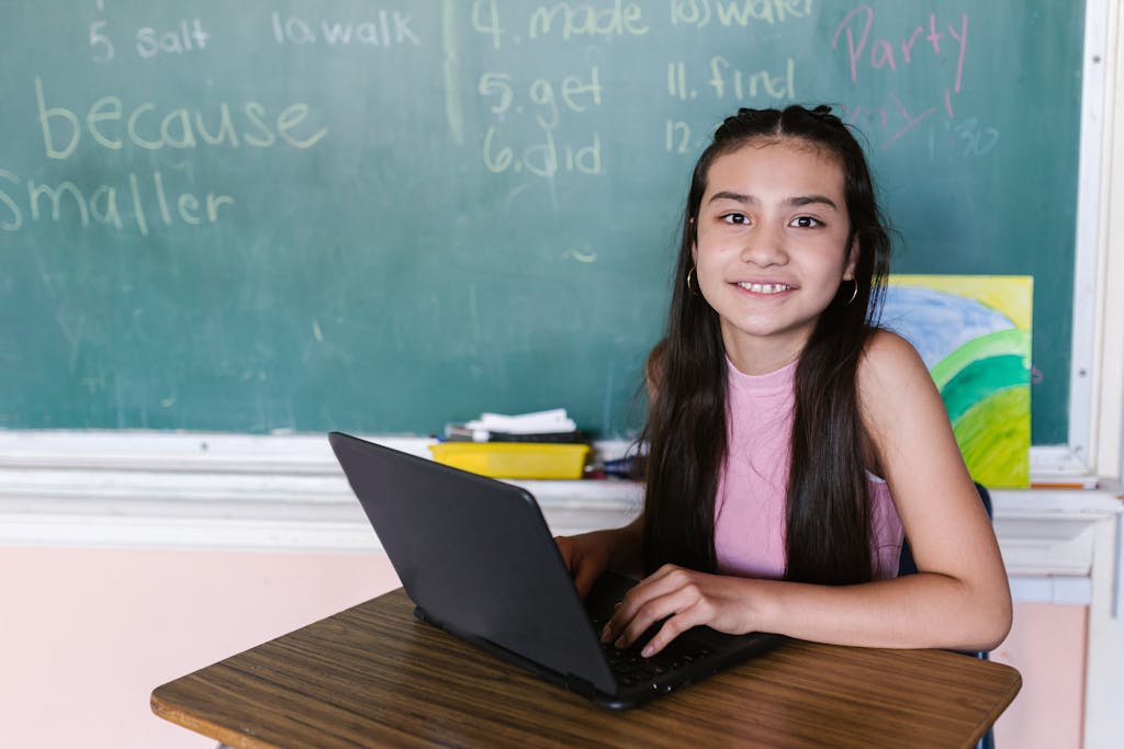 Young girl in a classroom, smiling while using a laptop, against a chalkboard backdrop.